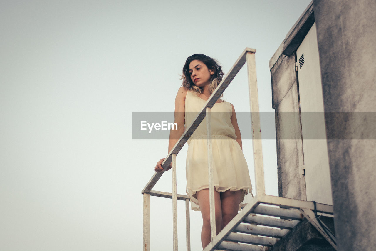 Low angle view of thoughtful young woman looking away while standing on building terrace during sunset