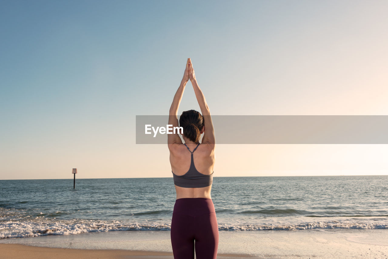 Back view of unrecognizable female in activewear standing in mountain pose with arms up while practicing yoga on sandy beach near waving sea