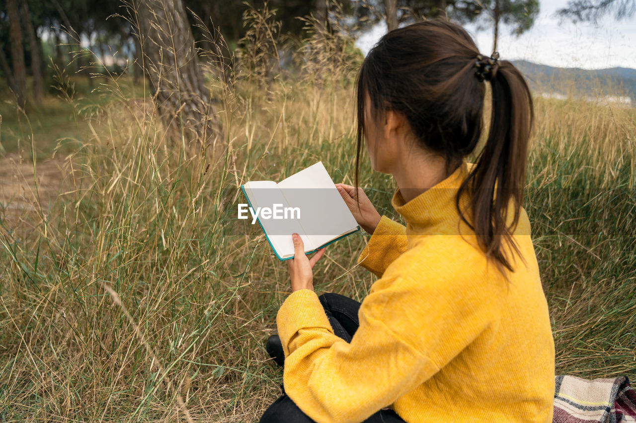 Side view of unrecognizable female sitting in filed in autumn and enjoying interesting story in book