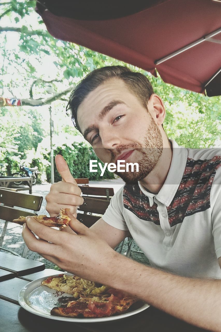 Portrait of young man gesturing thumbs up while eating pizza at restaurant