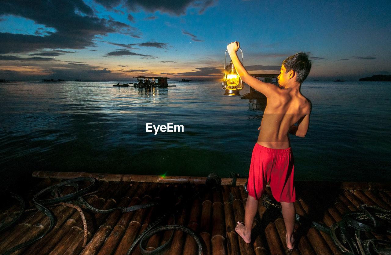 Rear view of shirtless boy holding oil lamp on wooden raft in sea at night