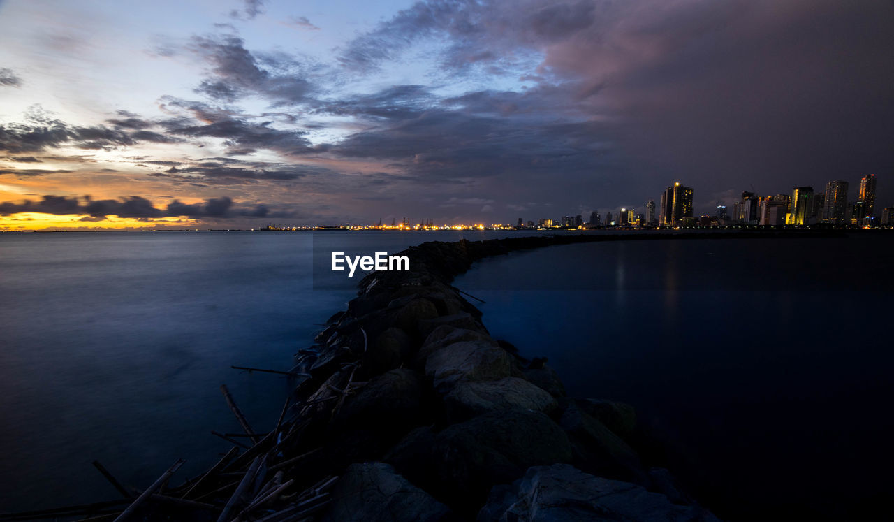 Sea by illuminated buildings against sky at sunset