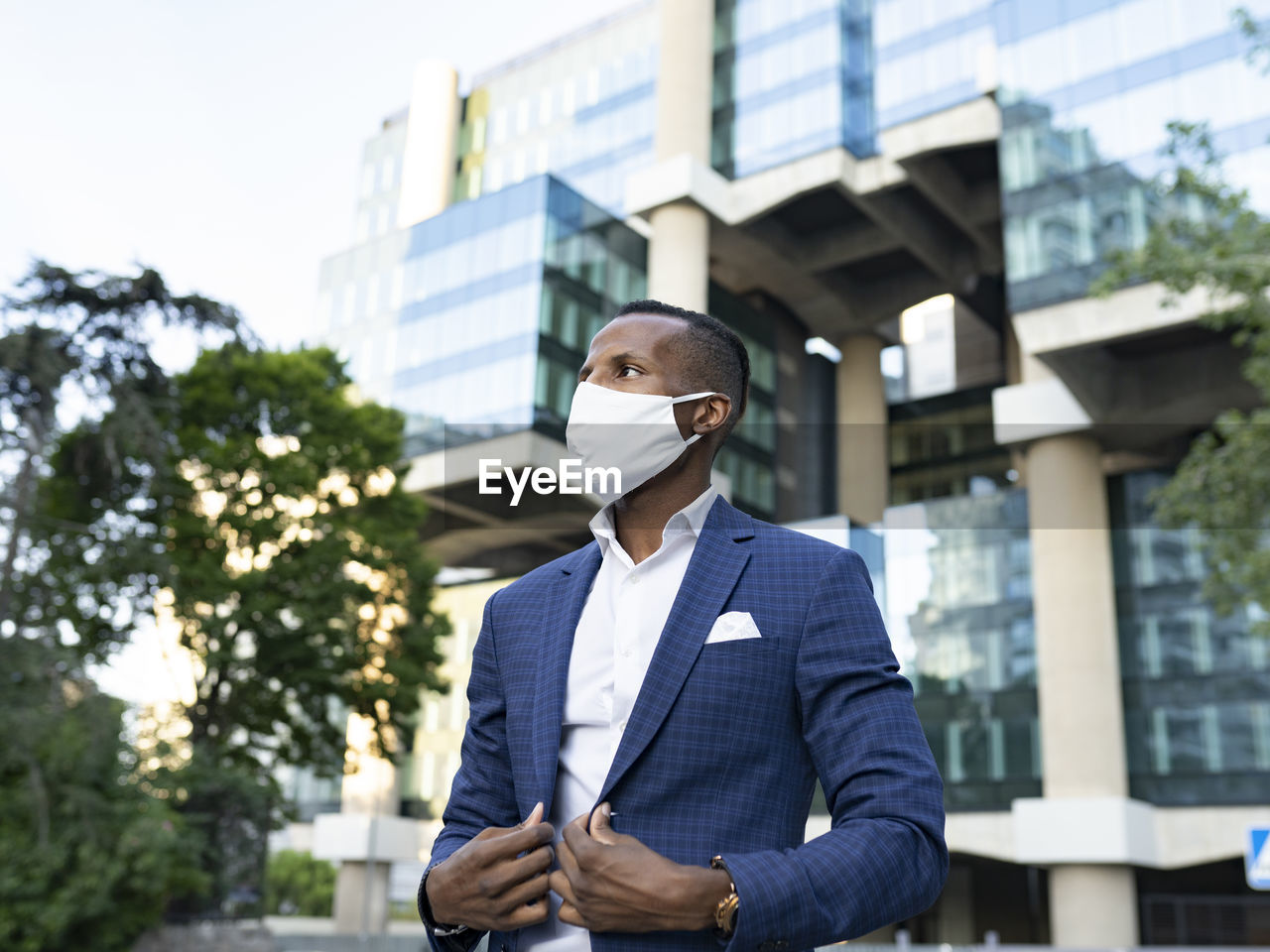 Serious black male entrepreneur in formal suit and medical mask standing in downtown waiting for meeting looking away