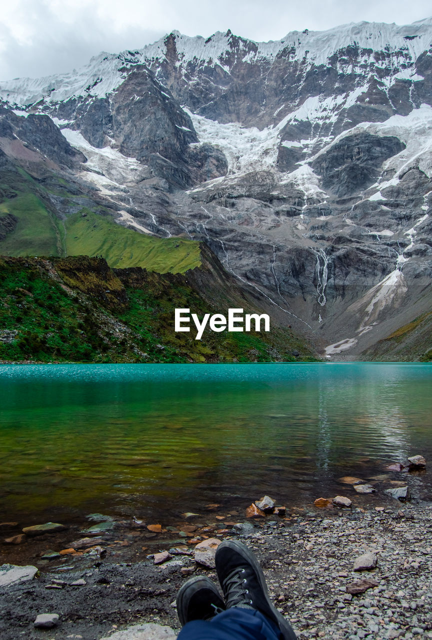 Low section of person resting by lake against snowcapped mountain