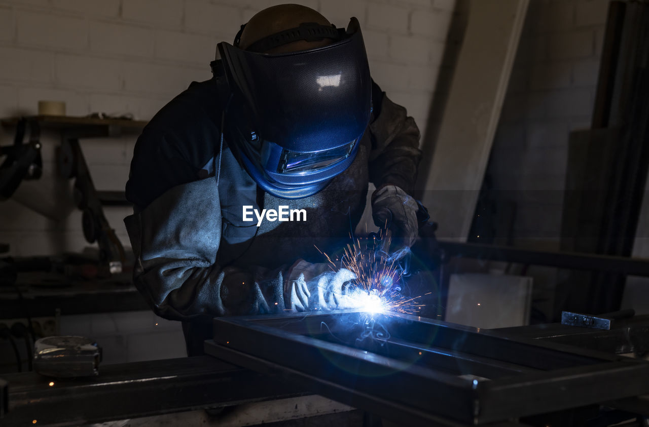 Unrecognizable male employee in protective gloves and helmet using welding machine while working in dark workshop