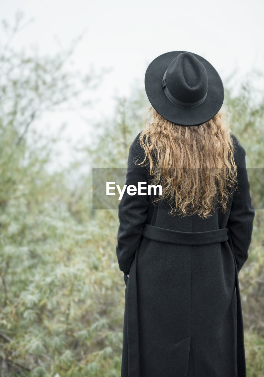 Portrait of young attractive woman in black coat and hat. autumn landscape, dry grass. back view