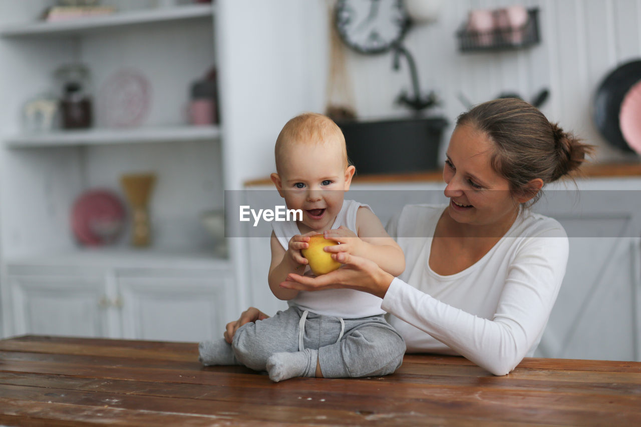 baby, child, childhood, indoors, person, parent, domestic life, two people, family, lifestyles, clock, togetherness, toddler, adult, women, home interior, female, domestic room, one parent, food, food and drink, wellbeing, smiling, emotion, healthy eating, happiness, bonding, clothing, men, furniture, positive emotion, fruit, sitting, eating, casual clothing, innocence, holding, human face, domestic kitchen, full length, love, kitchen, babyhood, flooring, care, looking, table, baby clothing, portrait, cute, father