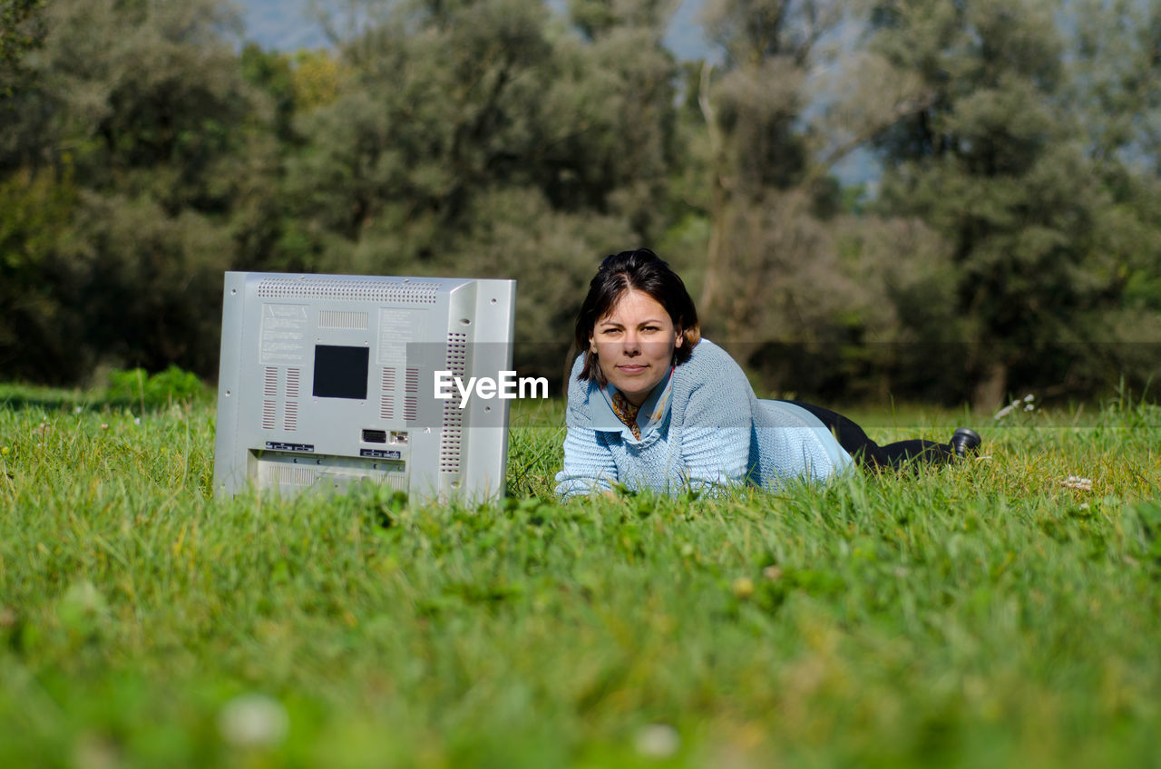 Portrait of woman lying with television set on grass during sunny day