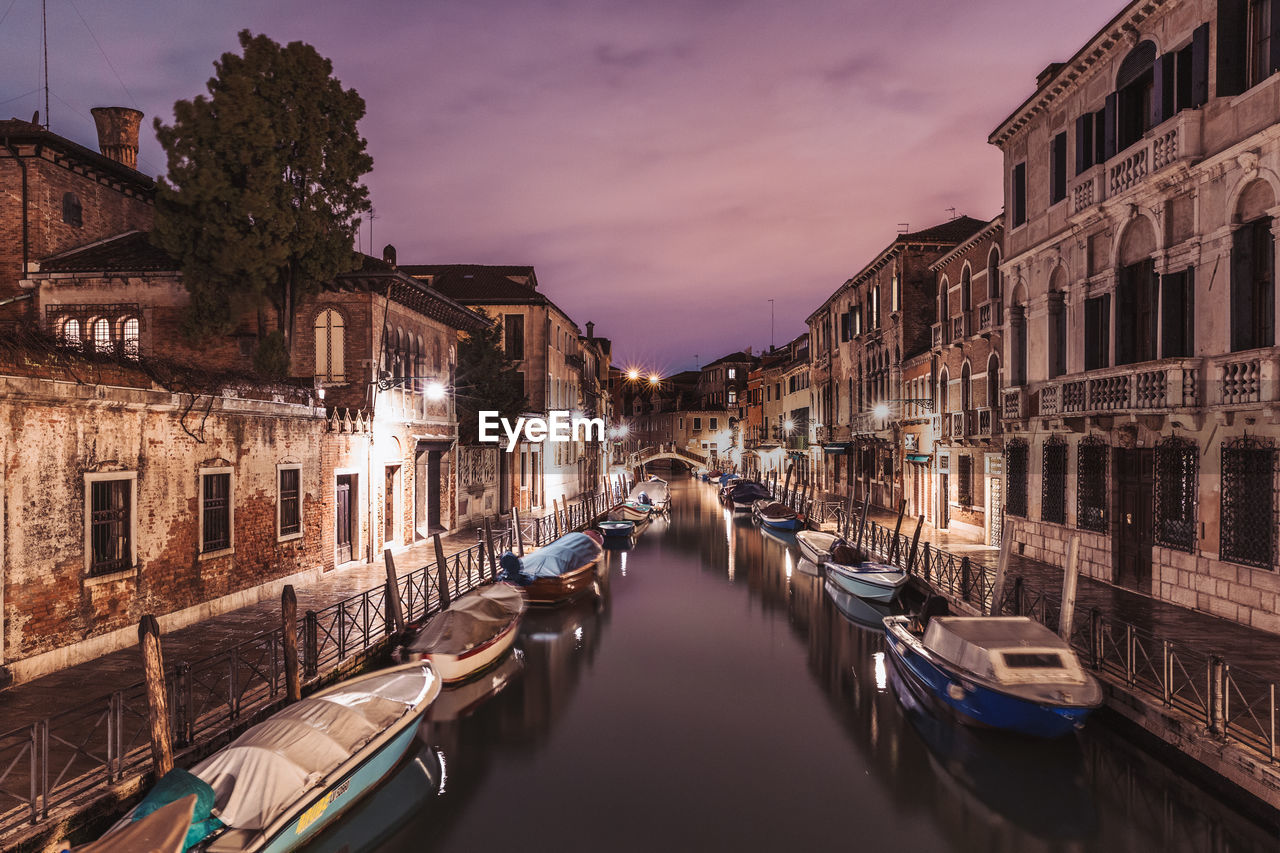 Boats moored on canal amidst illuminated buildings in city at night