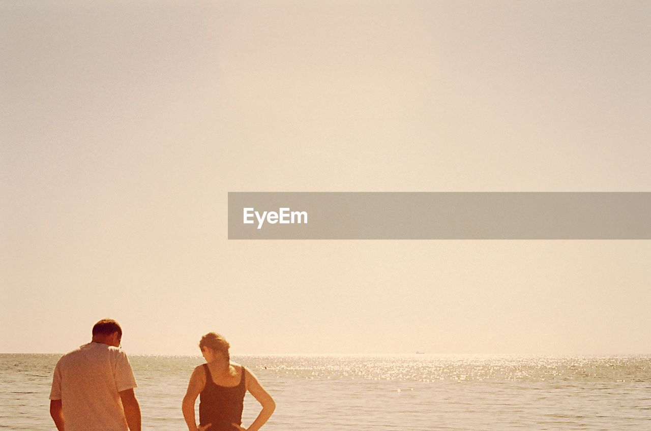 Couple standing at beach against clear sky on sunny day