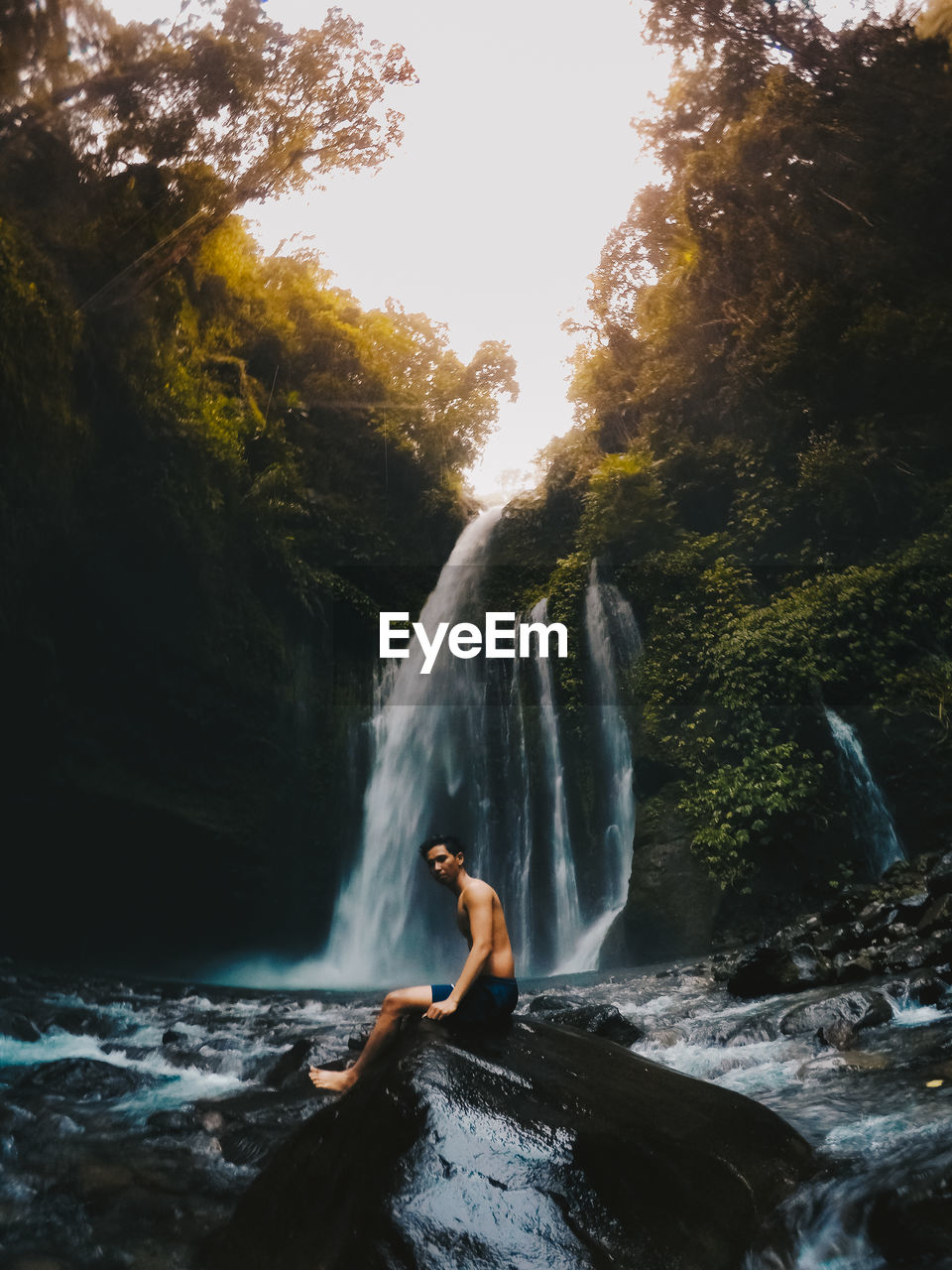 Side view of shirtless man standing by waterfall in forest