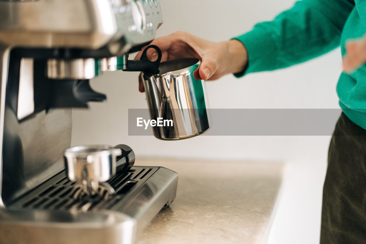 Crop unrecognizable person with metal jug preparing coffee with professional machine at table in house person