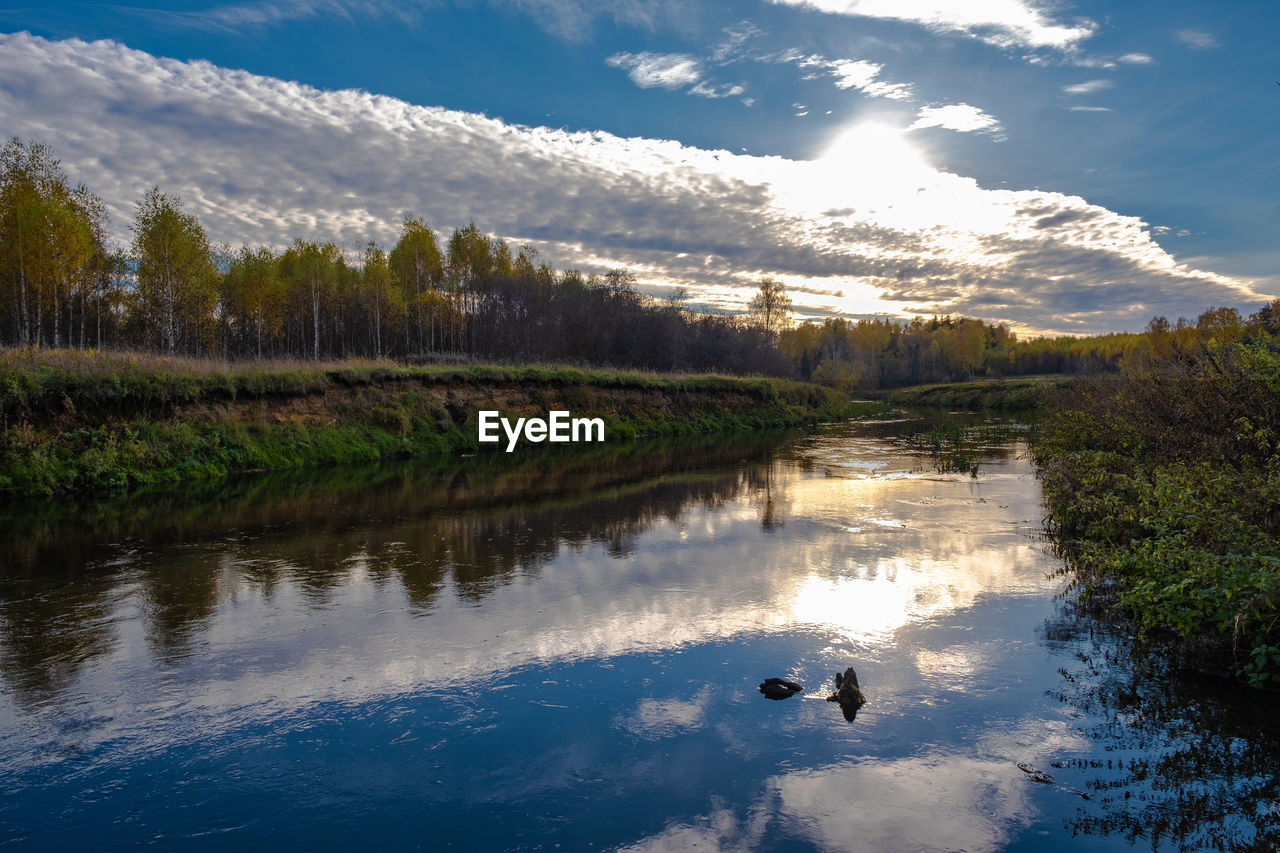 Reflection of the sky with clouds and the sun in the blue water of a small river, russia.