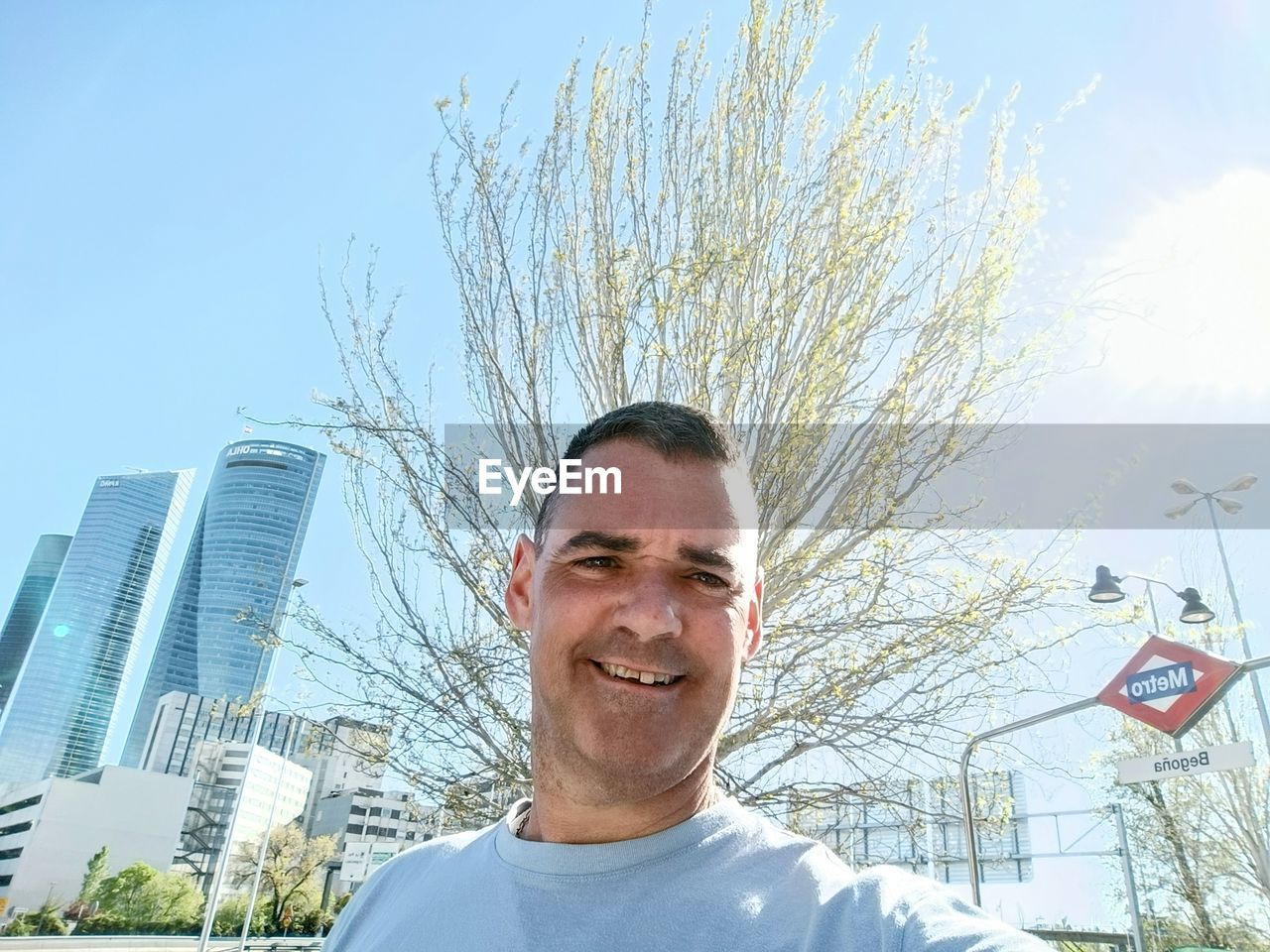 one person, portrait, smiling, adult, sky, men, architecture, happiness, headshot, city, building exterior, nature, day, built structure, front view, tree, emotion, office building exterior, sunlight, looking at camera, building, cheerful, skyscraper, standing, outdoors, low angle view, plant, blue, sunny, person, lifestyles, clear sky, smile, teeth, casual clothing, city life