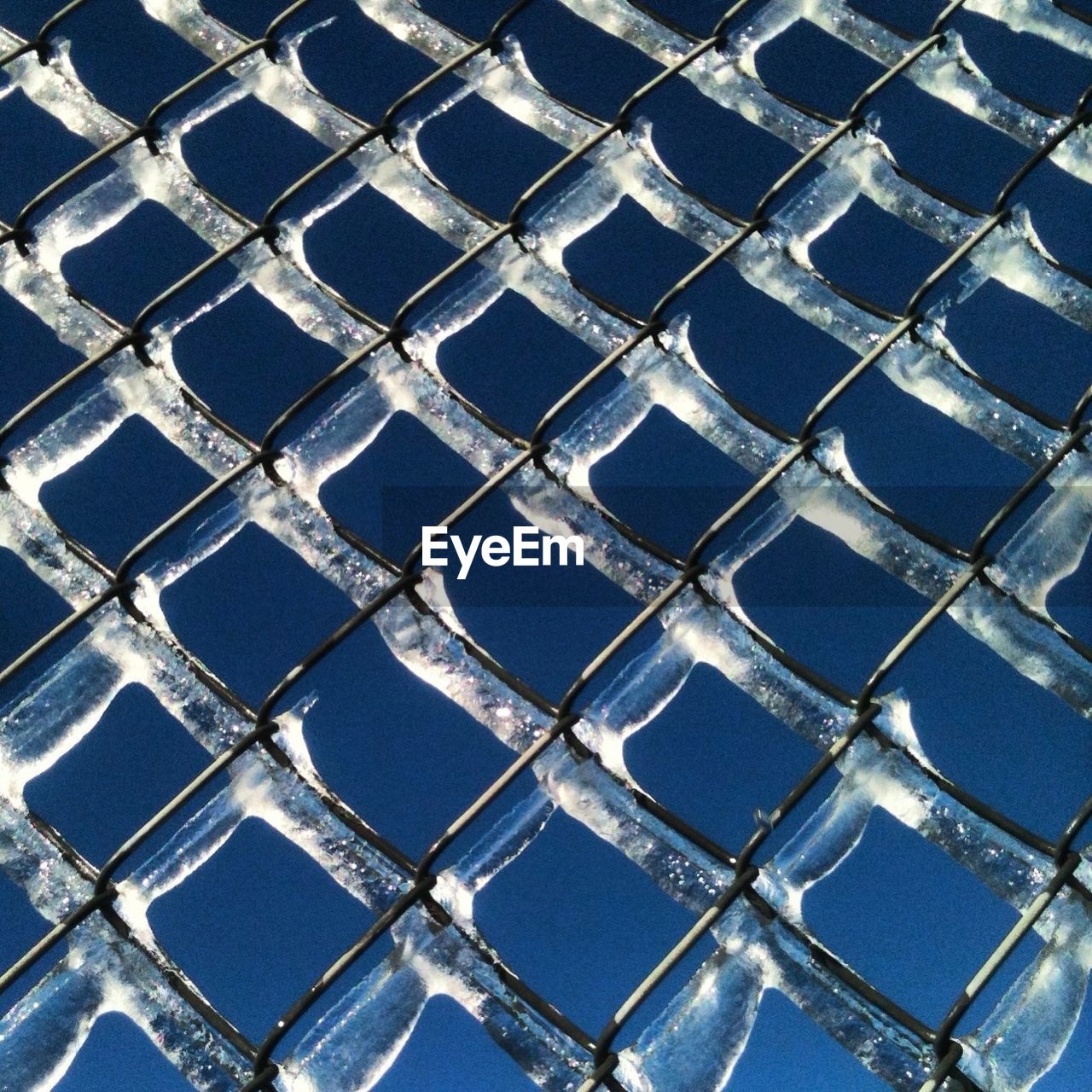 Full frame shot of chainlink fence covered with ice against blue sky