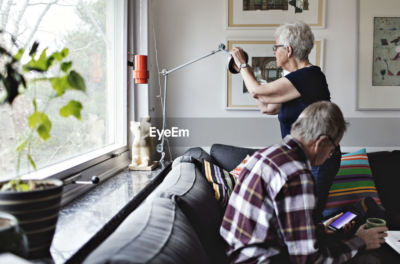 Senior woman adjusting floor lamp by man sitting on sofa in living room at home