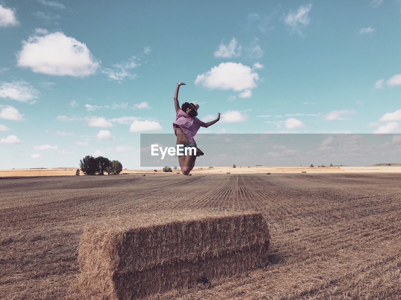 Woman with arms raised jumping over hay against sky