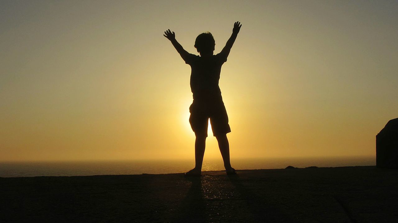 Full length of silhouette boy standing with arms raised on wall against sea during sunset