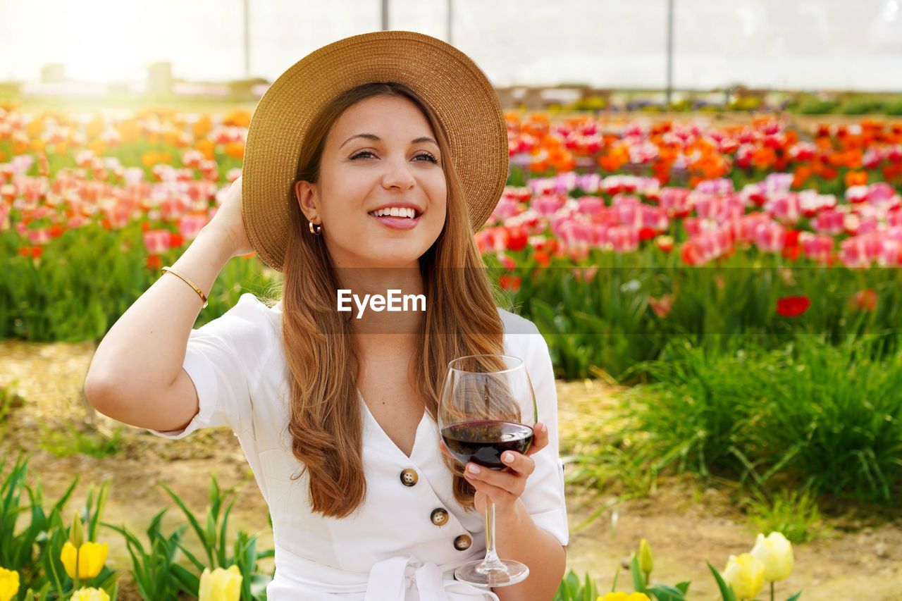 Woman drinking glass red wine over flowered background looking to the side with happy face smiling