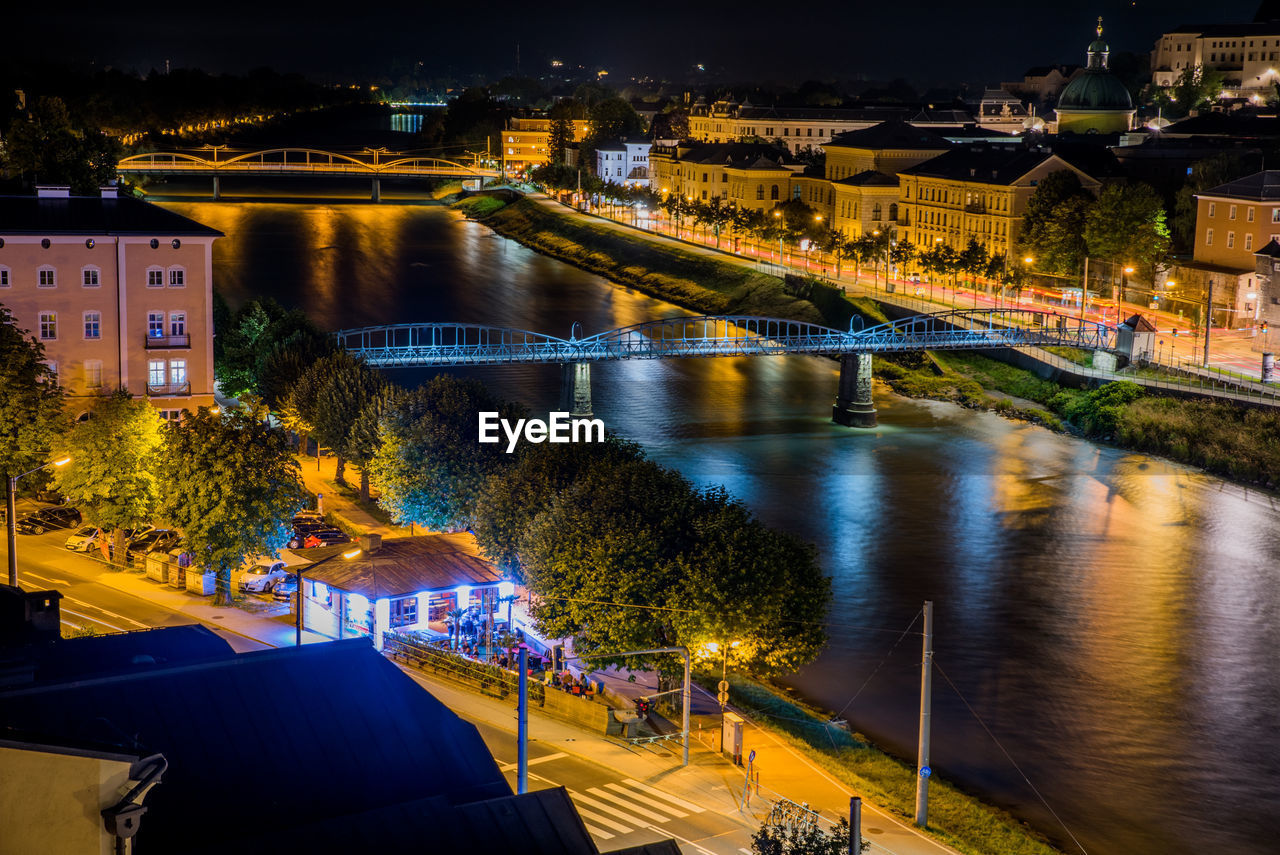 HIGH ANGLE VIEW OF ILLUMINATED BRIDGE OVER RIVER AT NIGHT