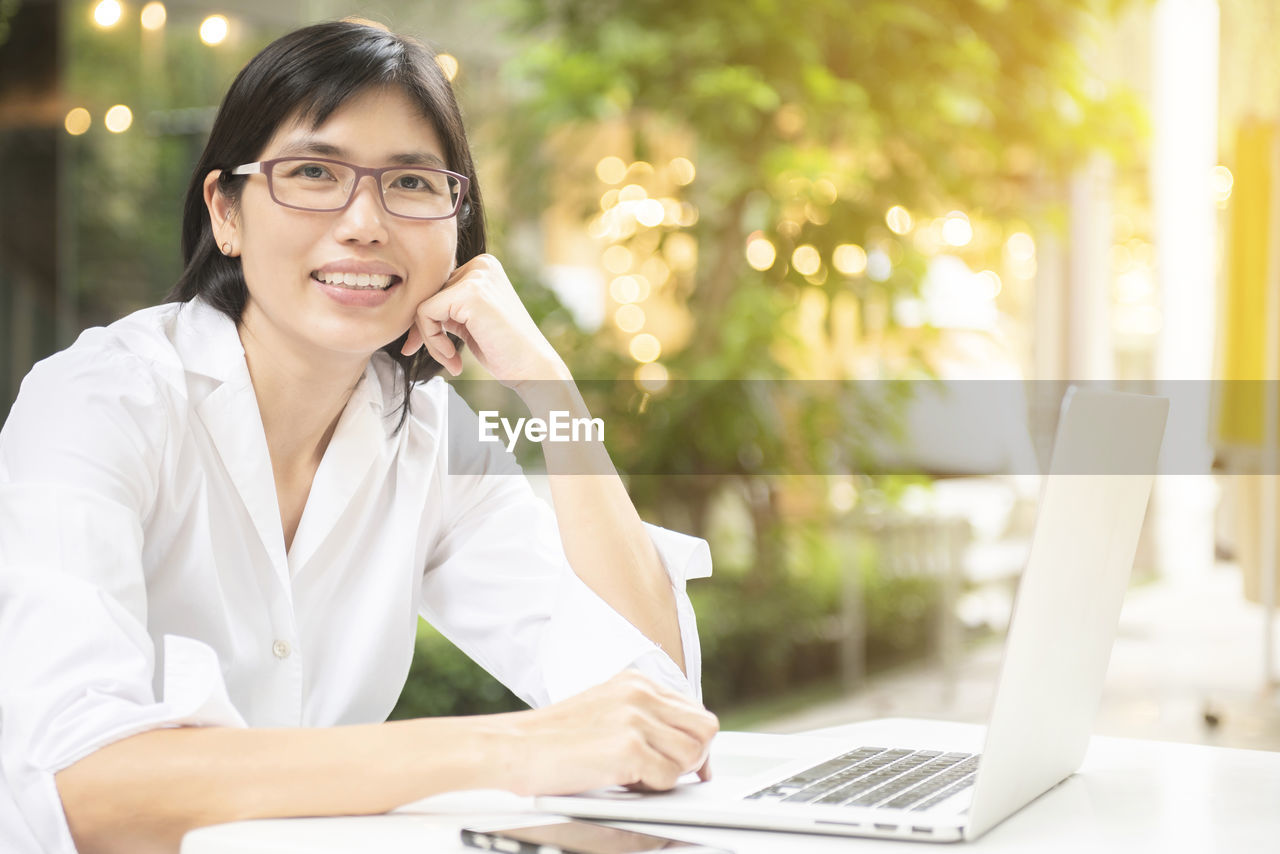 SMILING YOUNG WOMAN USING PHONE WHILE SITTING ON TABLE