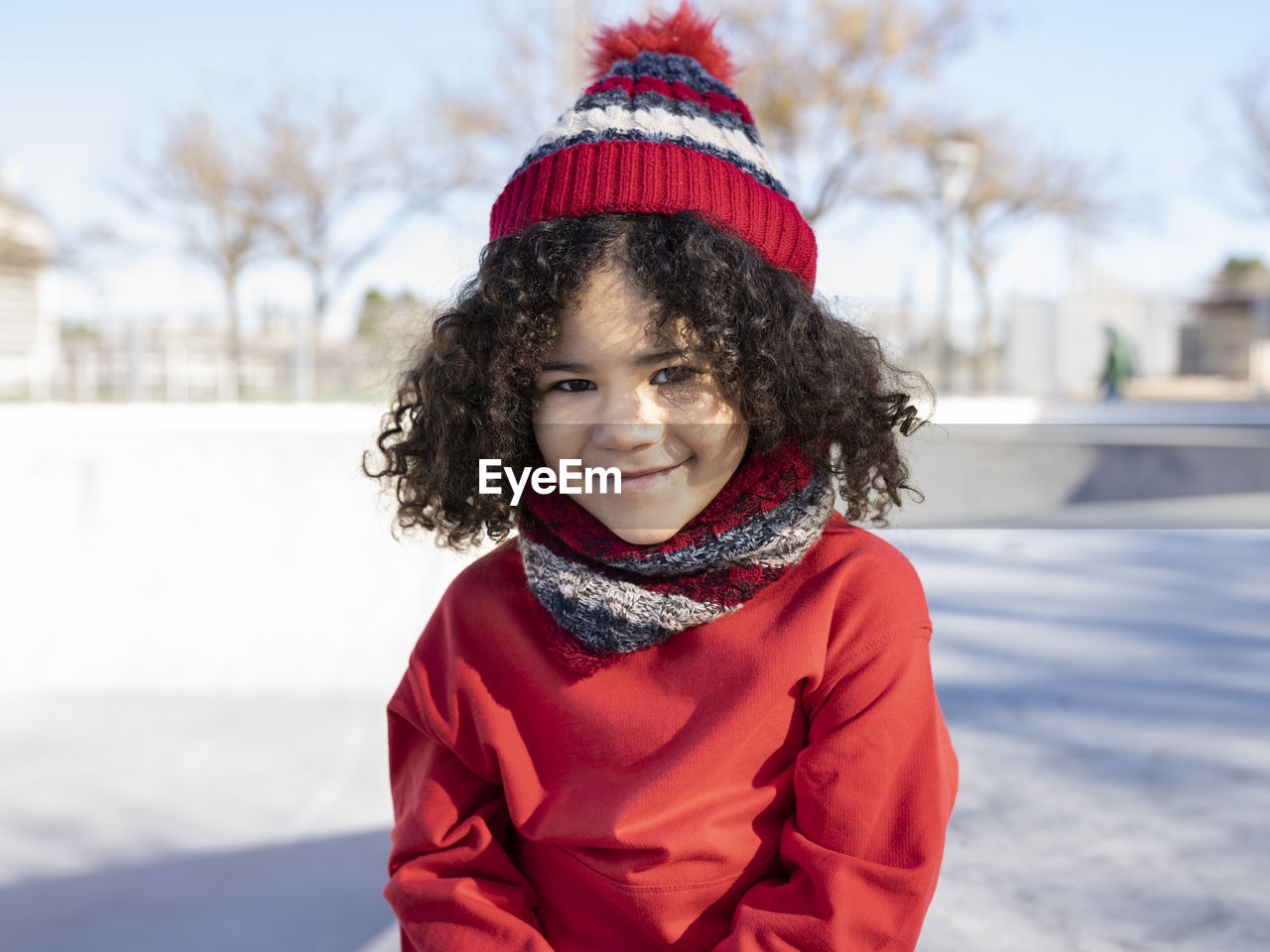 Smiling ethnic kid in warm beanie hat sitting on ramp in skate park and looking at camera