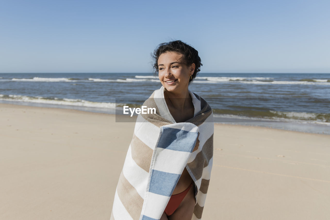 Smiling woman wrapped in towel looking away while standing at beach