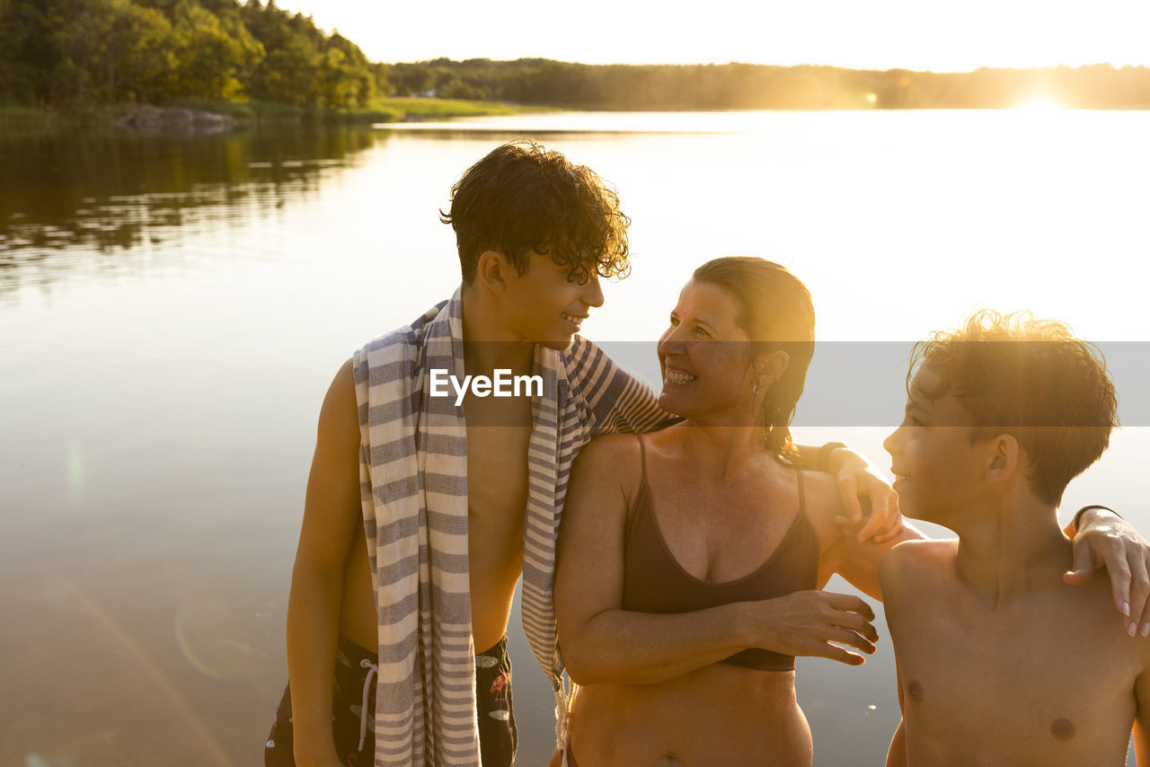 Happy sons talking with mother while standing against lake during sunset