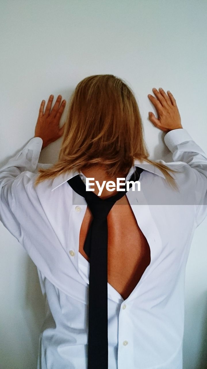 Rear view of woman wearing shirt and tie standing against wall at home