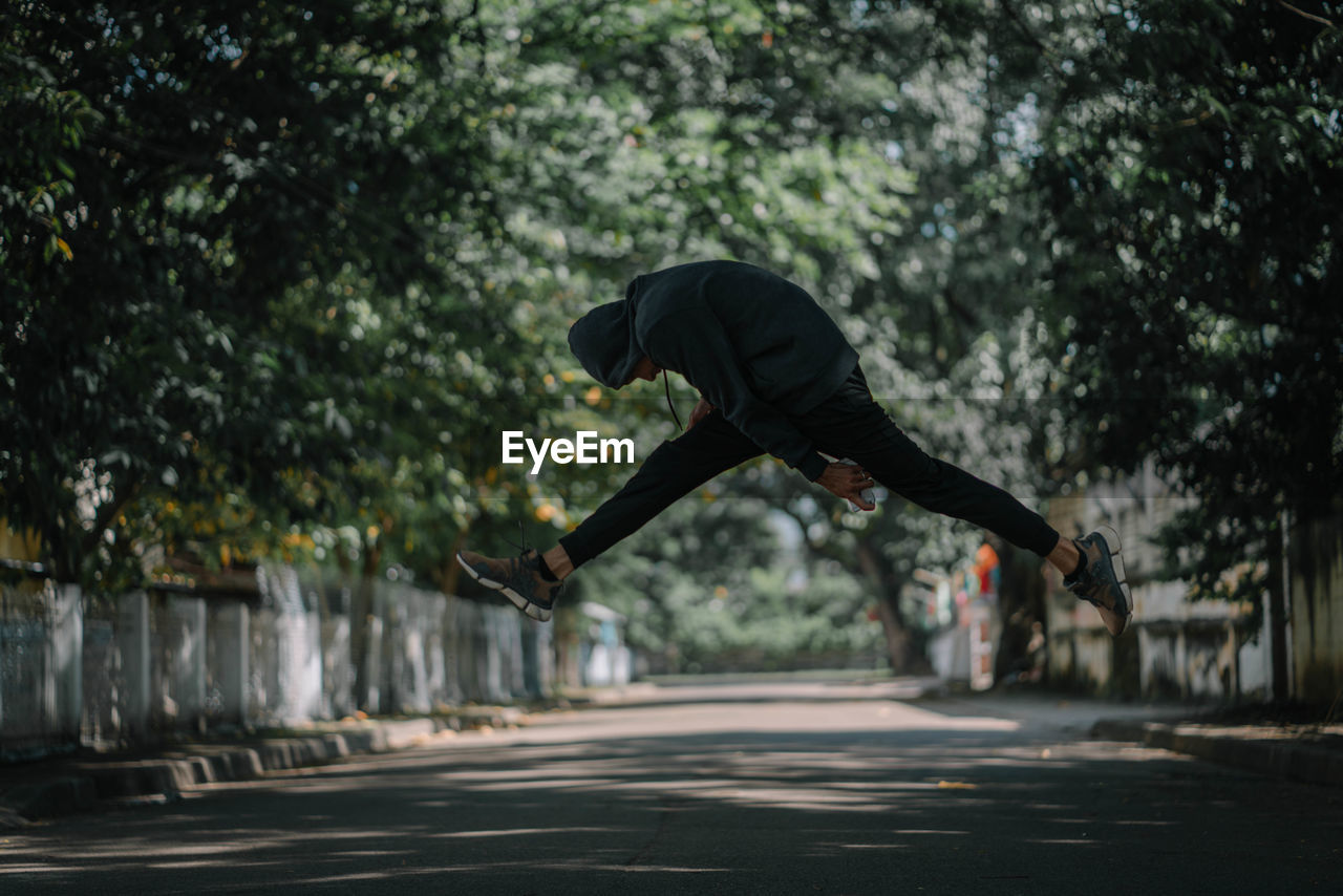Side view of young man jumping on road against trees