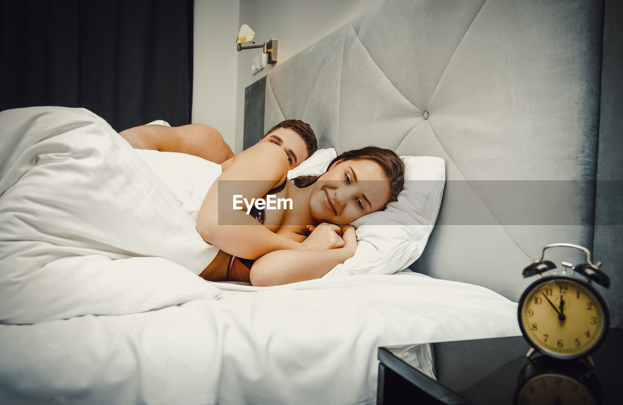 Young couple lying on bed with alarm clock on side table at home