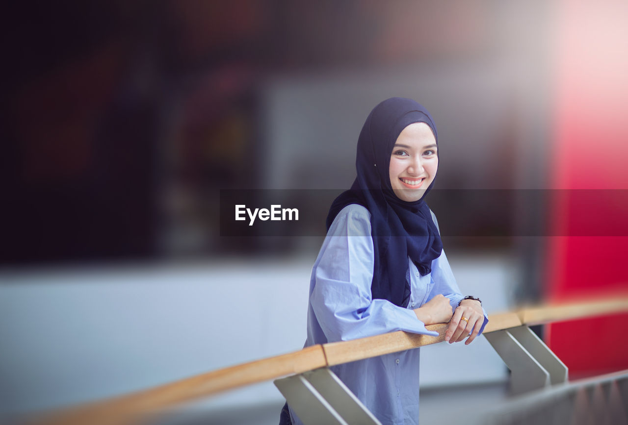 Portrait of teenage girl in hijab standing by railing in corridor