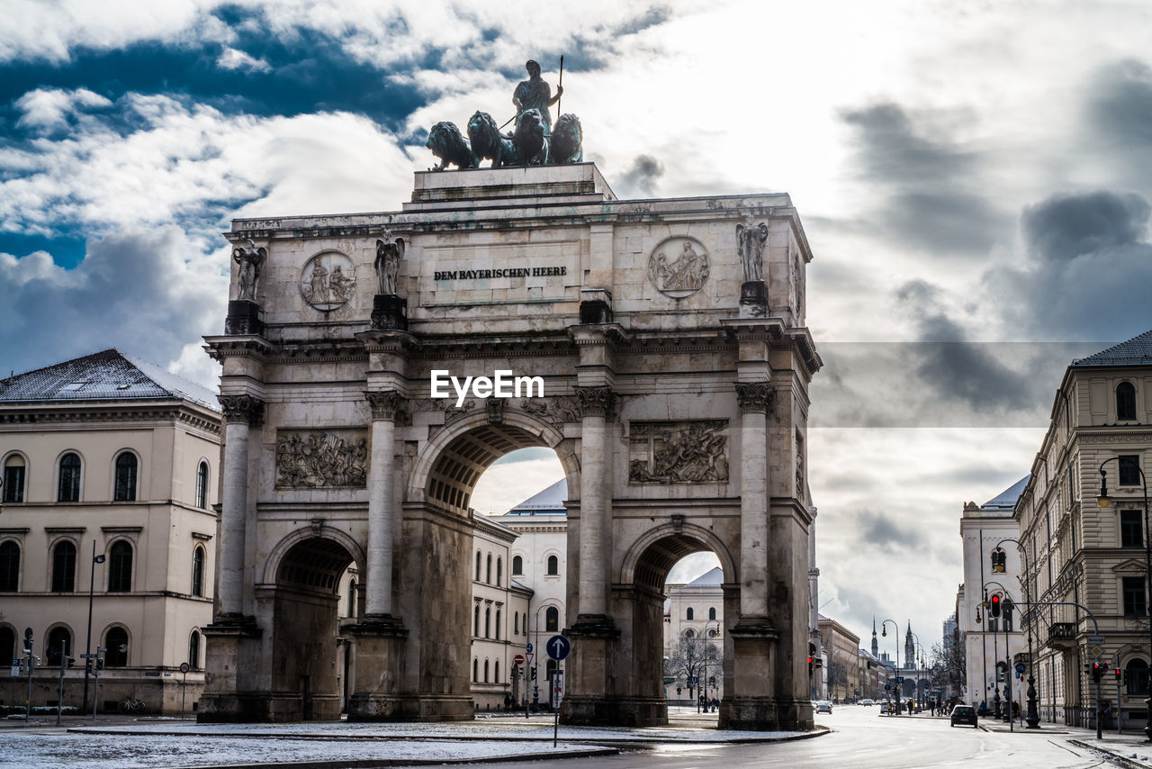 Low angle view of siegestor with buildings against cloudy sky