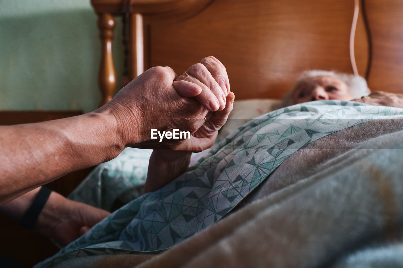 Cropped unrecognizable nurse holding hand of aged female patient with gray hair lying on bed in bedroom at home