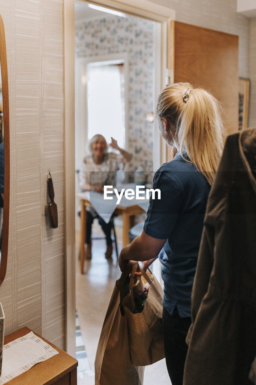 Female caregiver with bags looking at senior woman sitting at home