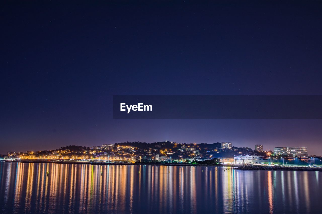 Illuminated city by sea against sky at night