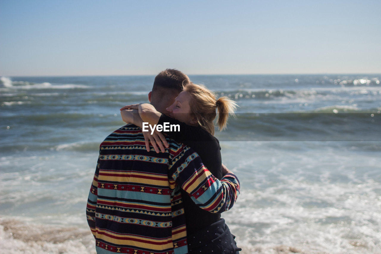 Couple embracing while standing at beach against sky