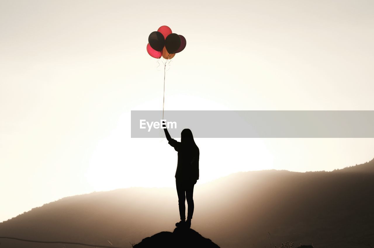 Full length of silhouette woman holding colorful balloons on cliff against mountain
