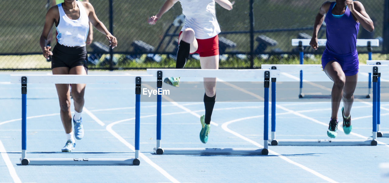 Three high school girls are racing in a 400 meter hurdle race on an outdoor track at a compeition.