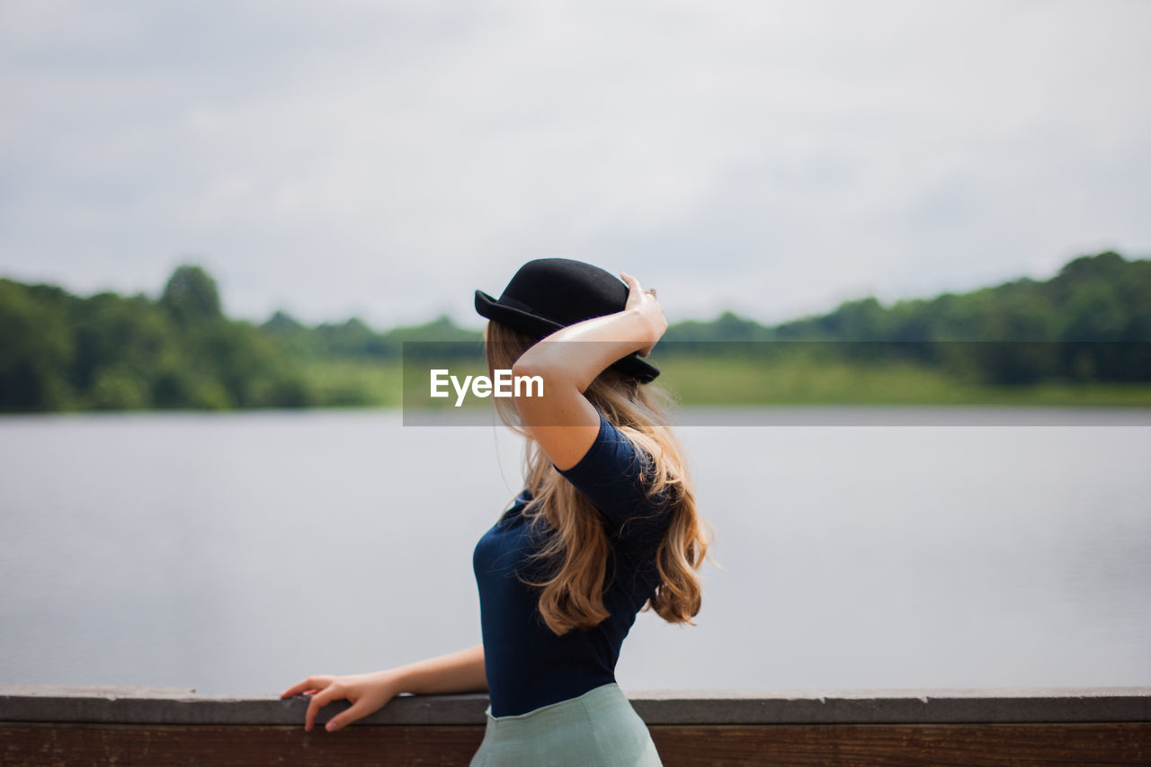 Woman wearing bowler hat standing by railing against lake