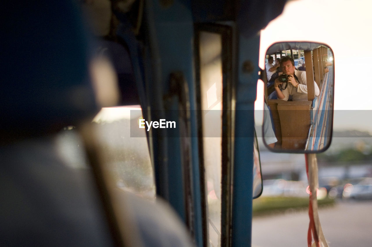 Man holding camera reflecting on bus side-view mirror