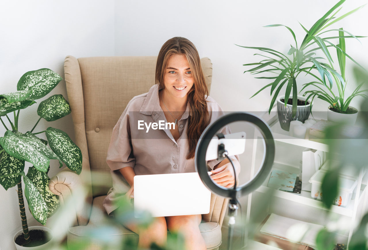 Young blogger in records video on mobile phone using ring lamp sitting on chair with laptop in room 