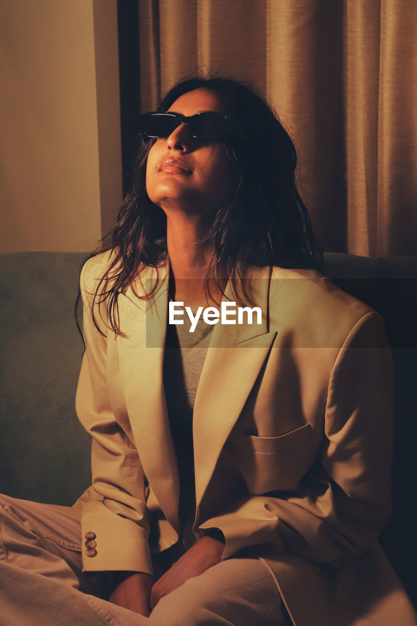 Portrait of young woman wearing sunglasses standing against curtains 