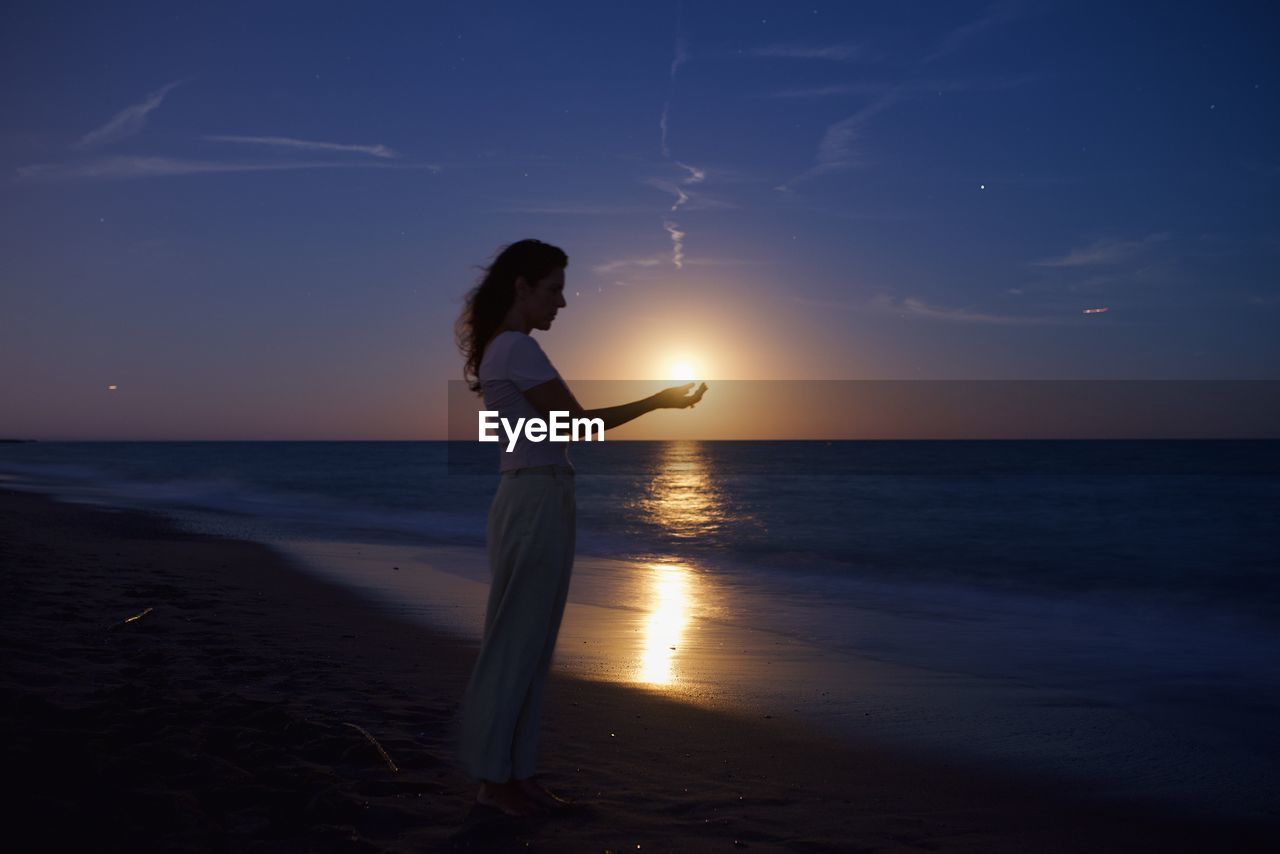 A woman on the beach holds the full moon in her hands