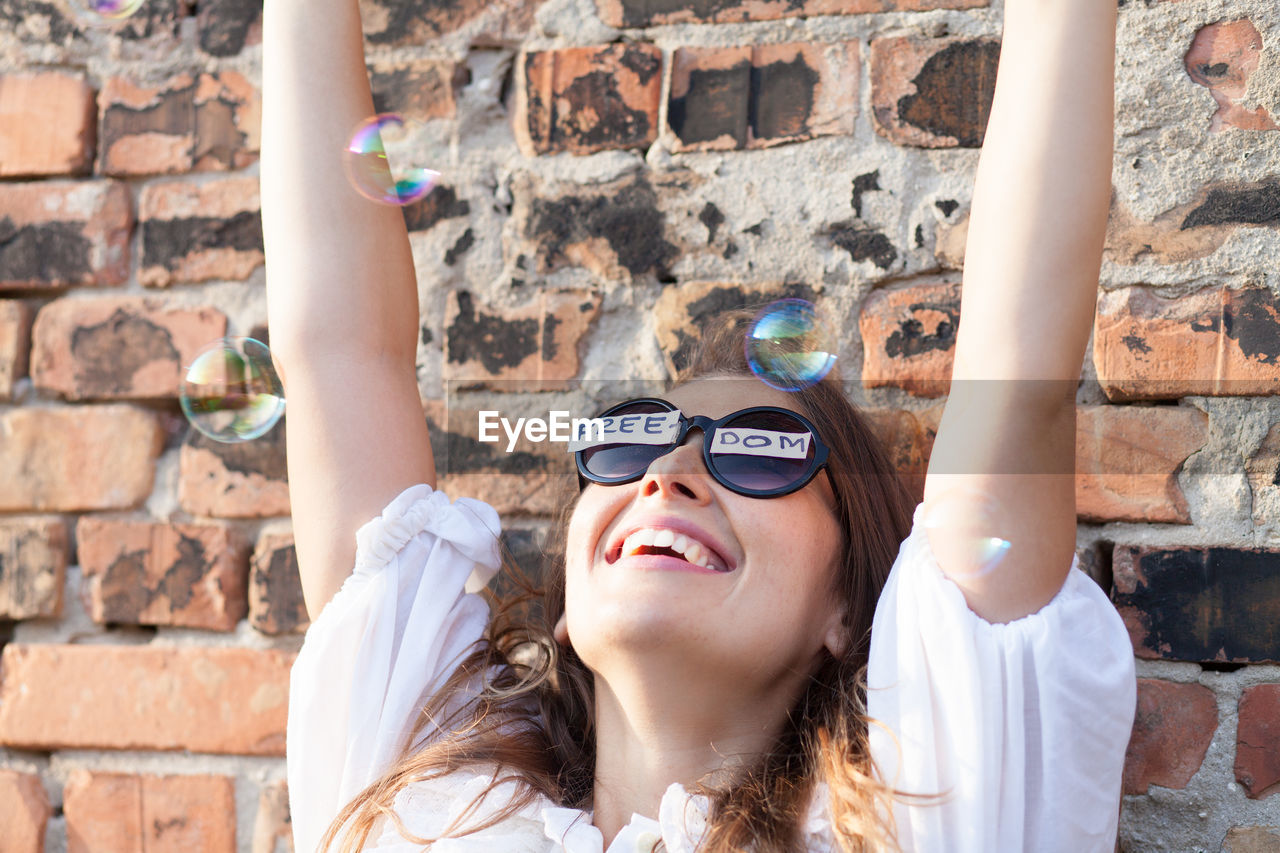 Cheerful woman wearing sunglasses with freedom text while playing with bubbles against brick wall