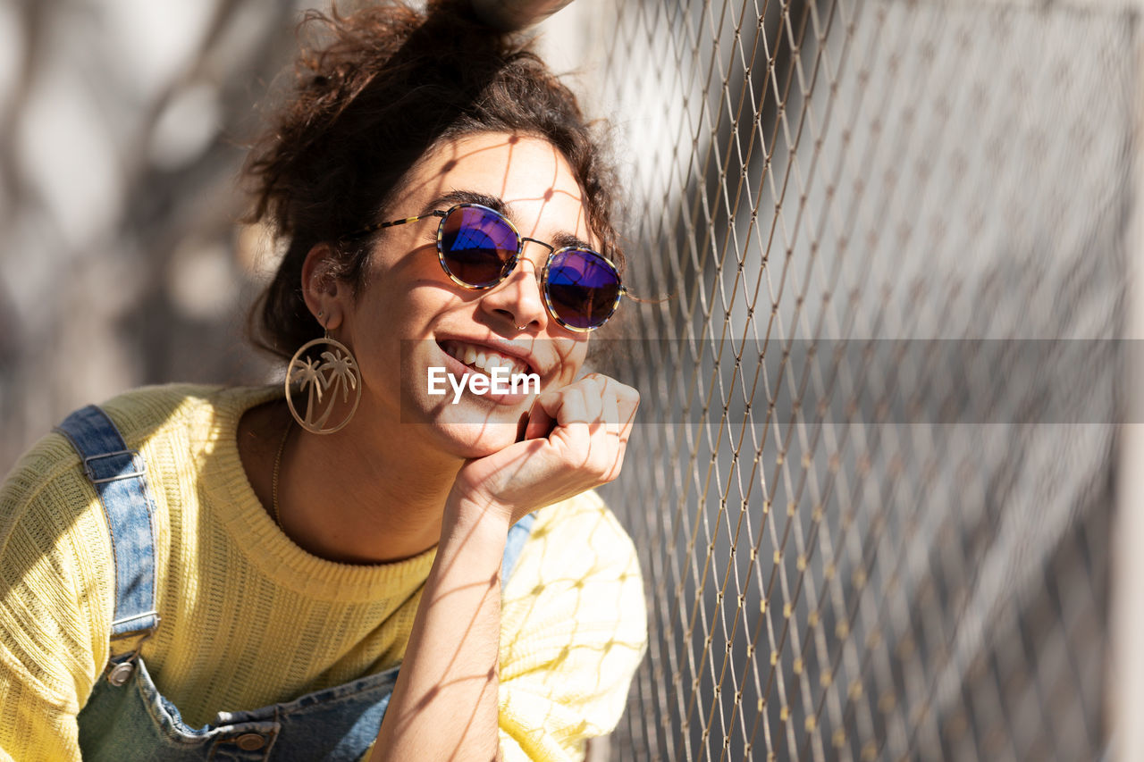 Modern cheerful millennial hispanic female with curly hair wearing yellow sweatshirt with denim overalls and trendy sunglasses and earrings sitting leaning on hand near mesh fence in sunlight
