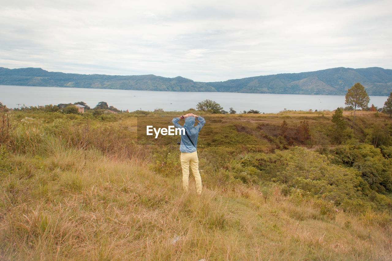 REAR VIEW OF WOMAN STANDING ON FIELD BY MOUNTAIN AGAINST SKY