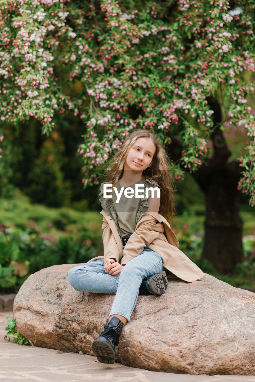 Cute teenage girl with long brown hair in a trench coat is sitting on a rock in the park