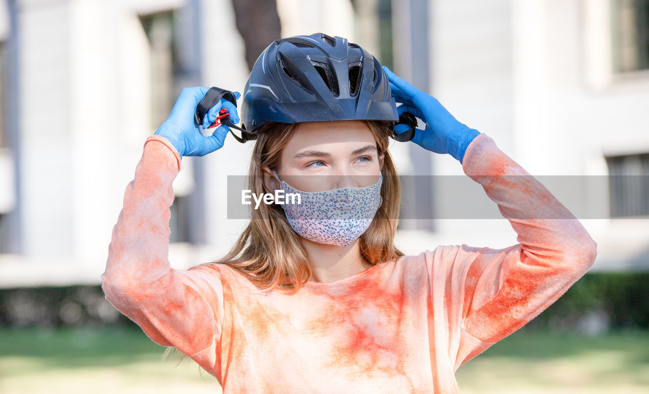 Young female cyclist in protective mask and gloves putting on hardhat and looking away on street of madrid, spain