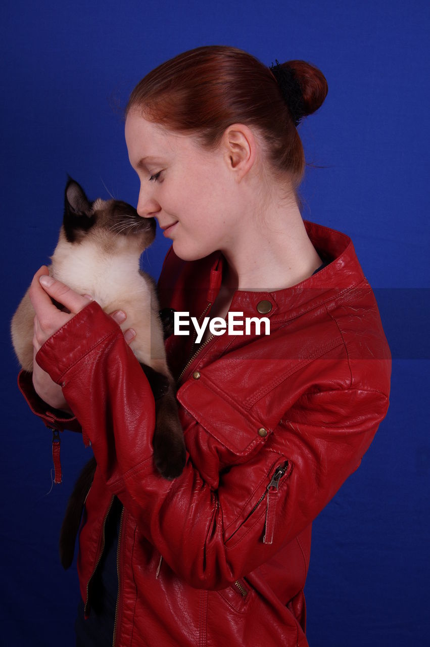 Young woman holding siamese cat against blue background